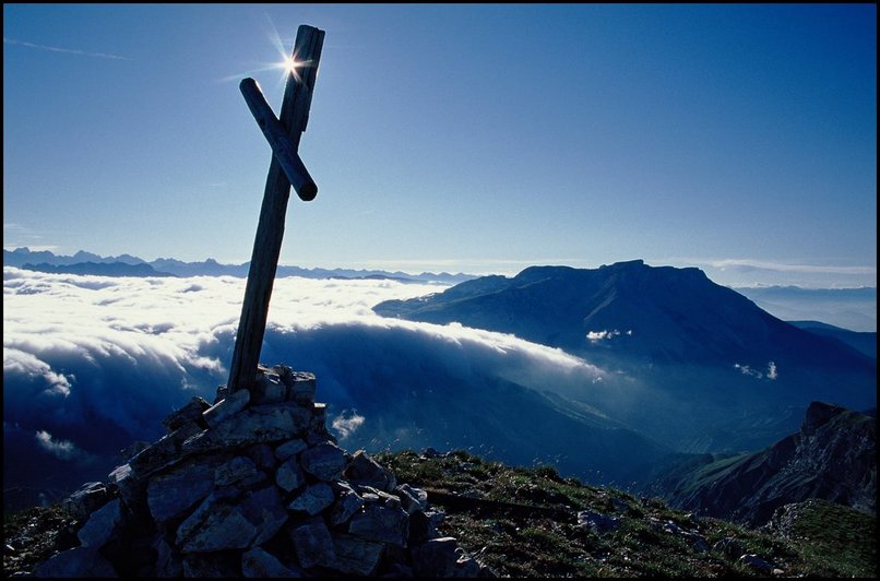 Photo : La petite croix, deux bouts de bois, qui marque le sommet de la Tête de Garnesier, en Dévoluy. Nuages bas sur l'intérieur du massif, et soleil au dessus. 
